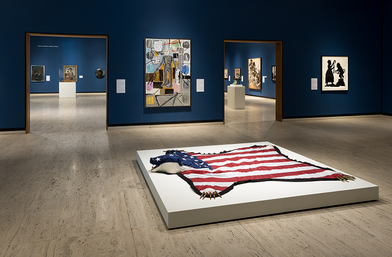 Art gallery with an American flag sculpture on a platform, surrounded by colorful artworks on blue walls.