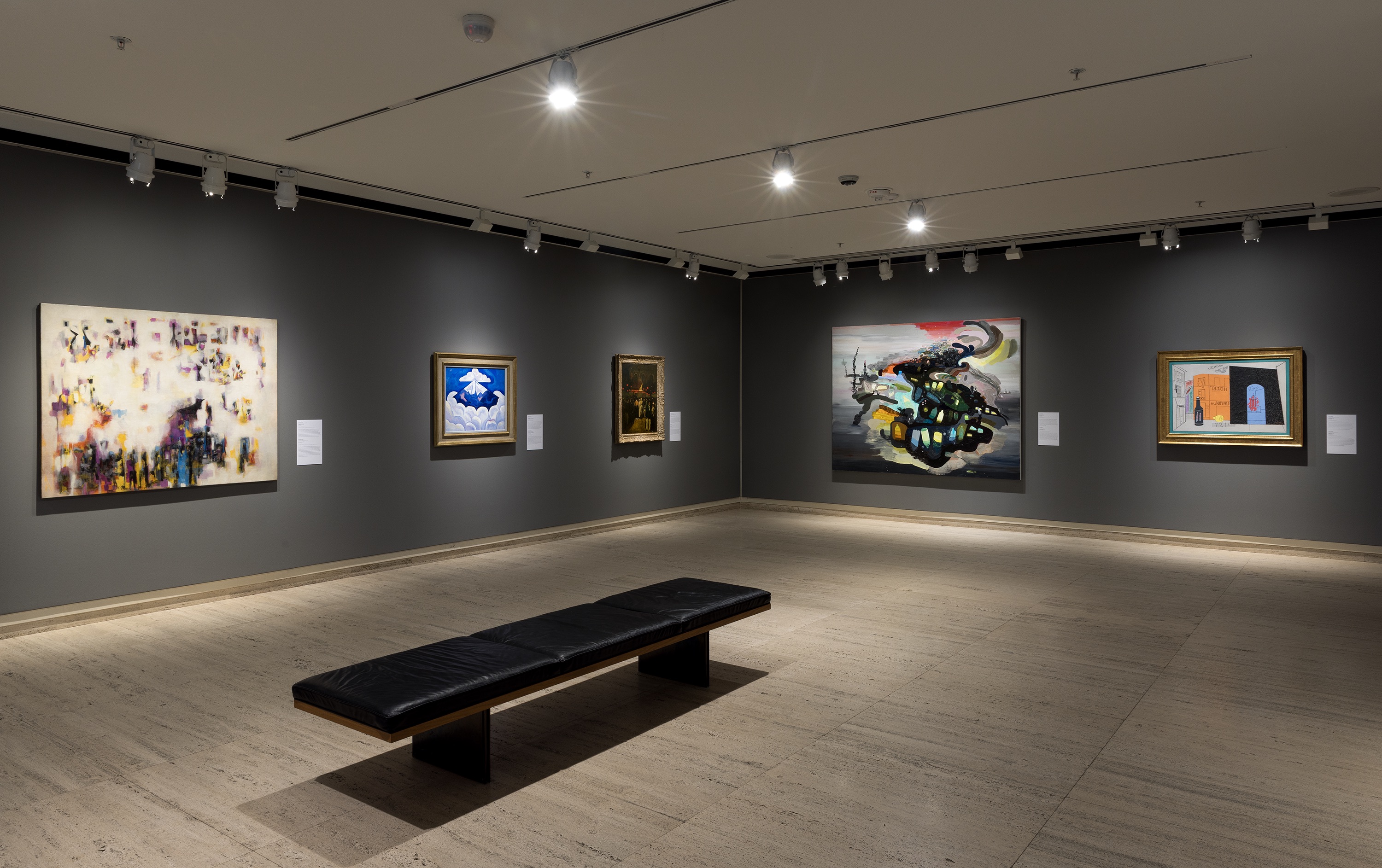 Art gallery with modern paintings on dark walls, a bench on light flooring, and bright overhead lighting.