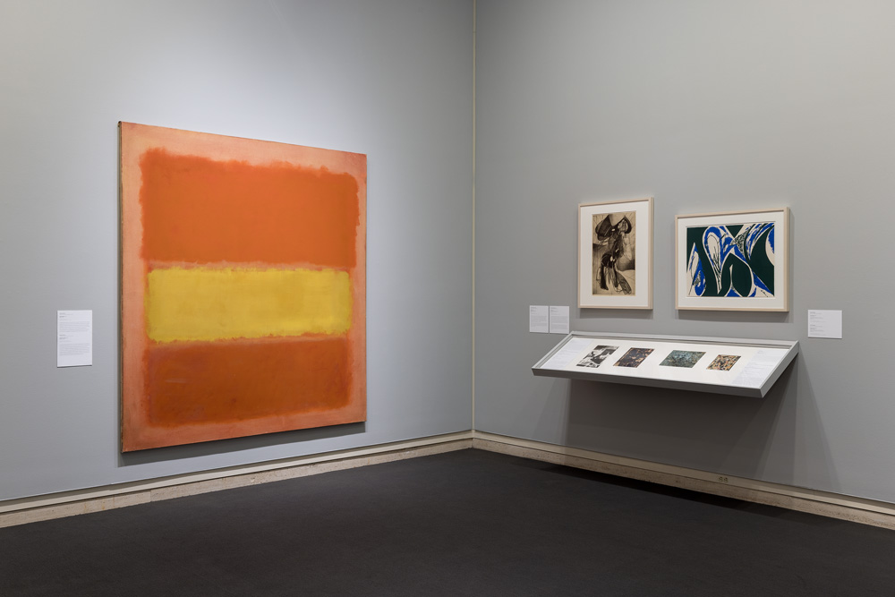 Large abstract orange and yellow painting on the left, framed artworks and display case on the right.