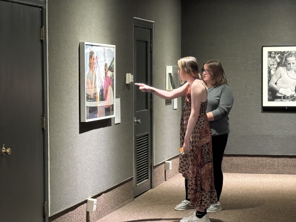 Two women viewing framed art in a dimly lit gallery.