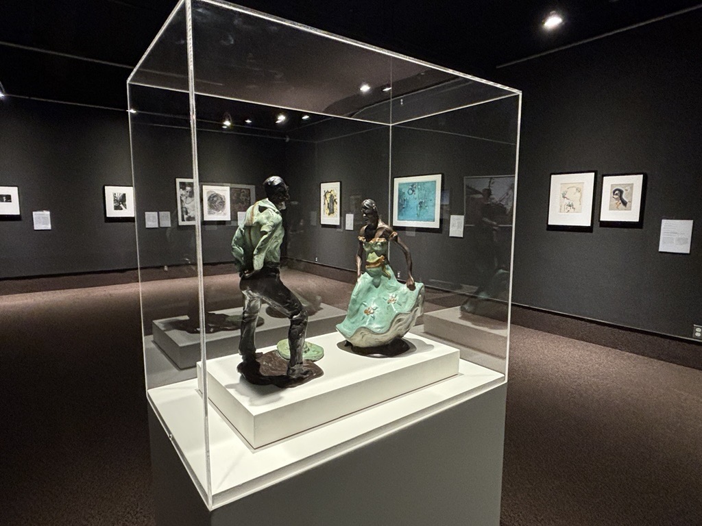 Sculptures of two figures in a glass case, surrounded by framed artwork in a dim gallery.