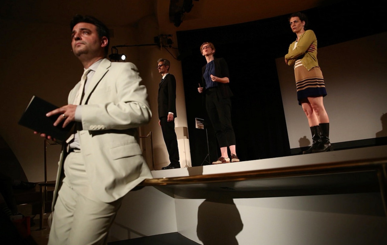 A man in a white suit holds a book, with three people standing on a stage in the background.