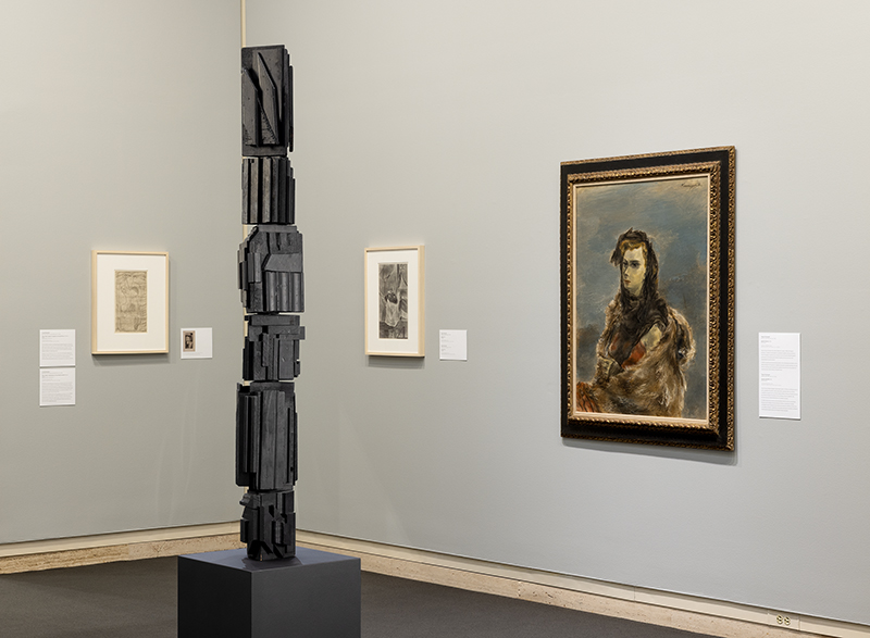 Gallery with abstract black sculpture, framed portrait, and artworks on beige walls.