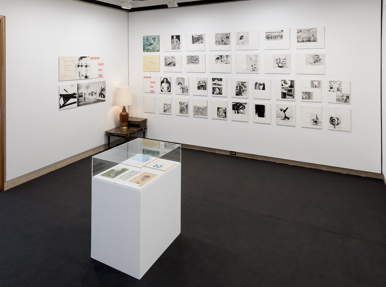 Gallery room with framed artwork on white walls, a pedestal display case, and a lamp on a side table.