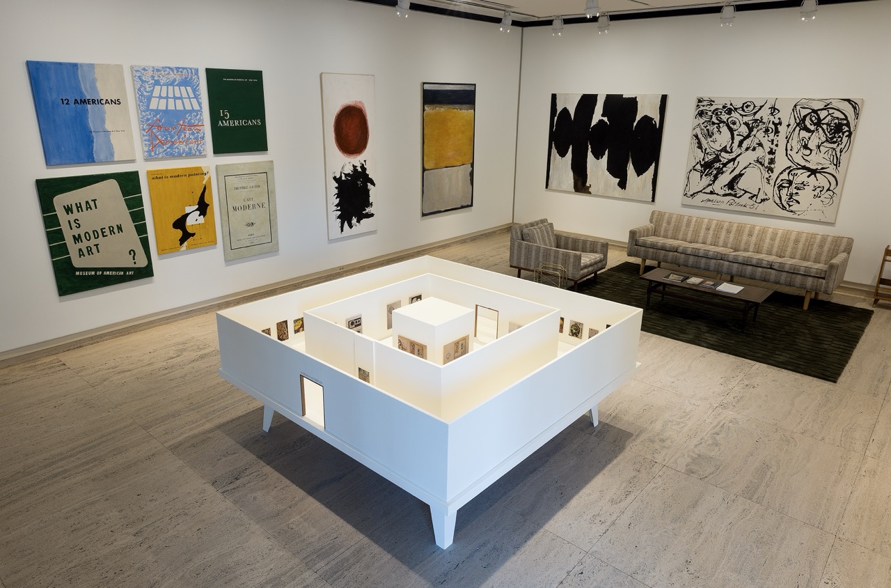 Modern art gallery with paintings, a scale model display, and gray couches on a rug.