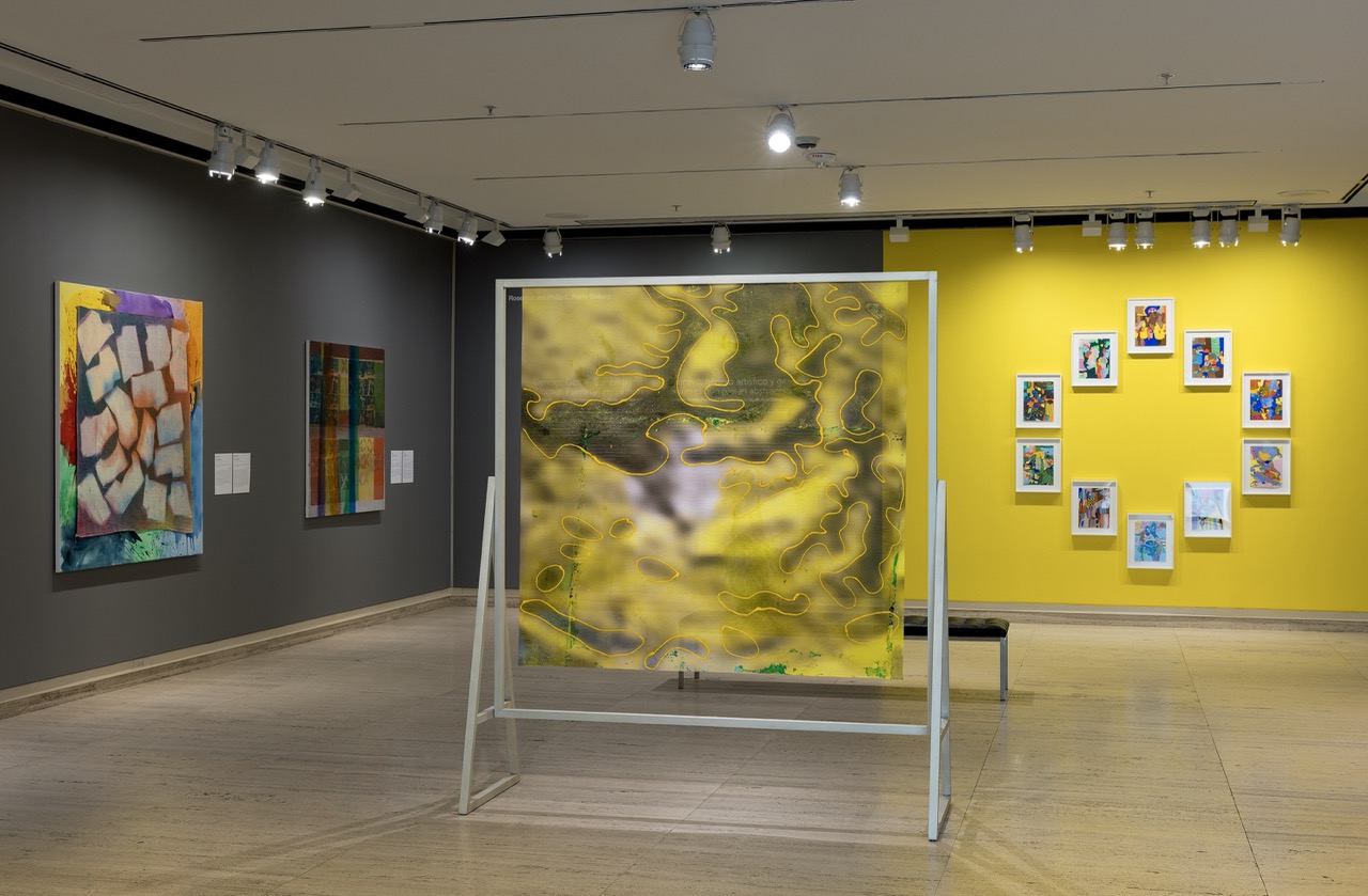 Gallery space with colorful abstract paintings on gray and yellow walls, and art behind glass frame.