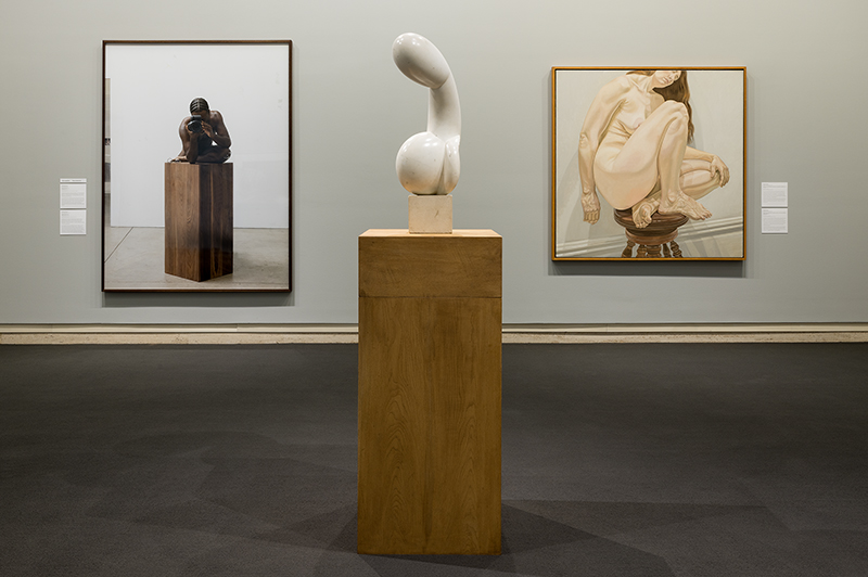 Sculpture displayed on wooden pedestal, flanked by two framed art pieces of seated nude figures.