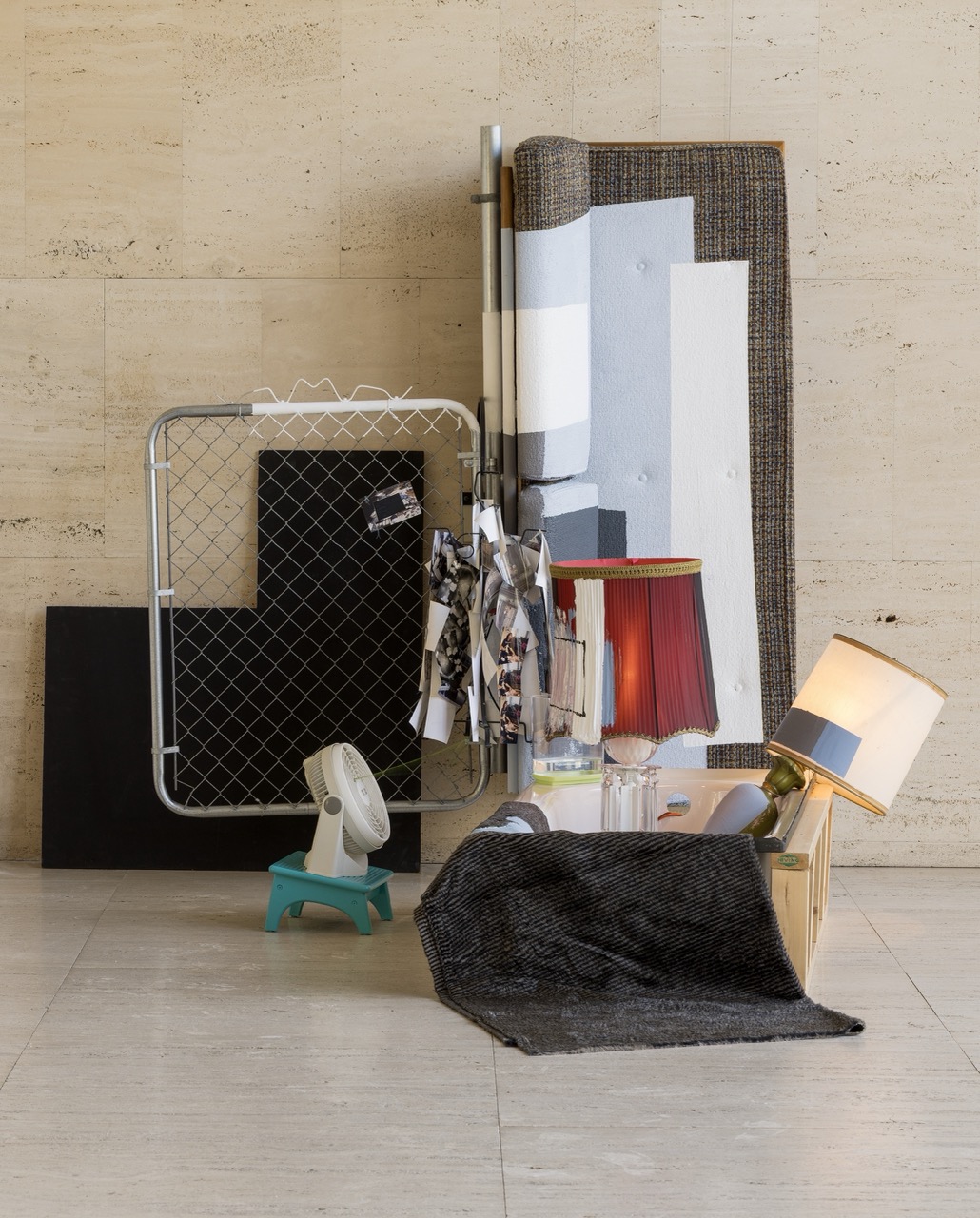 Abstract arrangement of furniture, fabric, and household items against a beige wall.