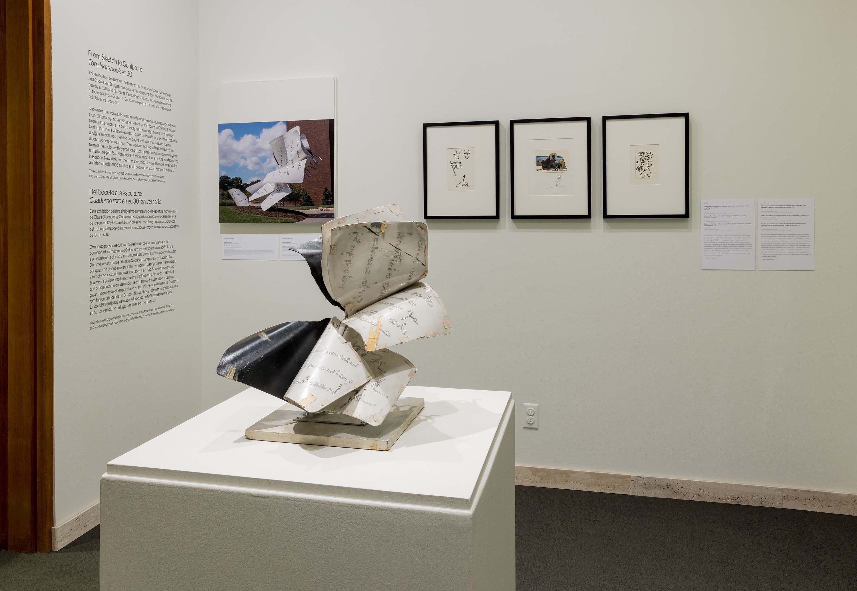 Sculpture with curved, layered forms on a pedestal, surrounded by framed art on a gallery wall.