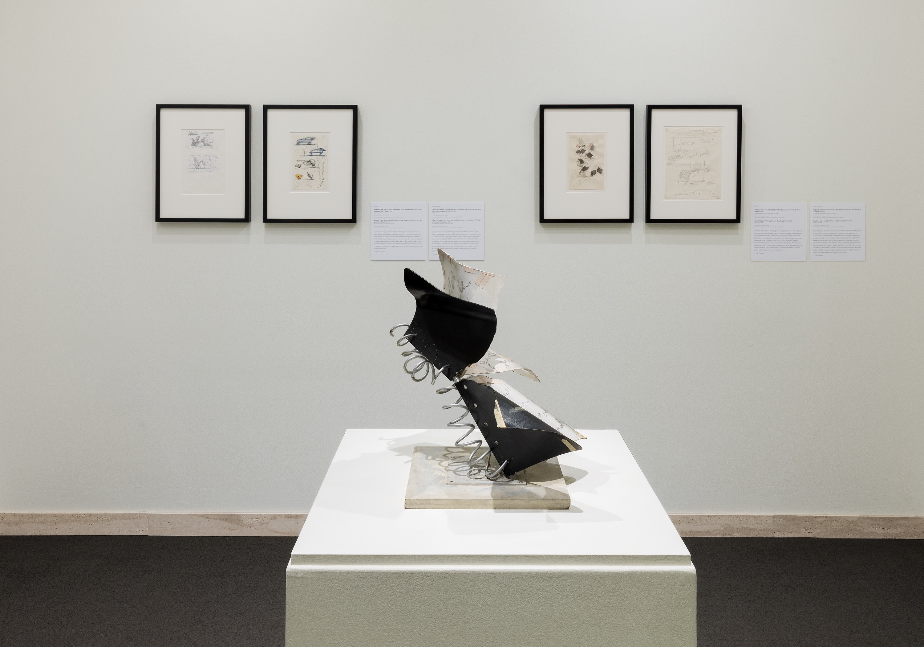 Sculpture with metal springs on a pedestal, framed artworks displayed on a white wall in the background.