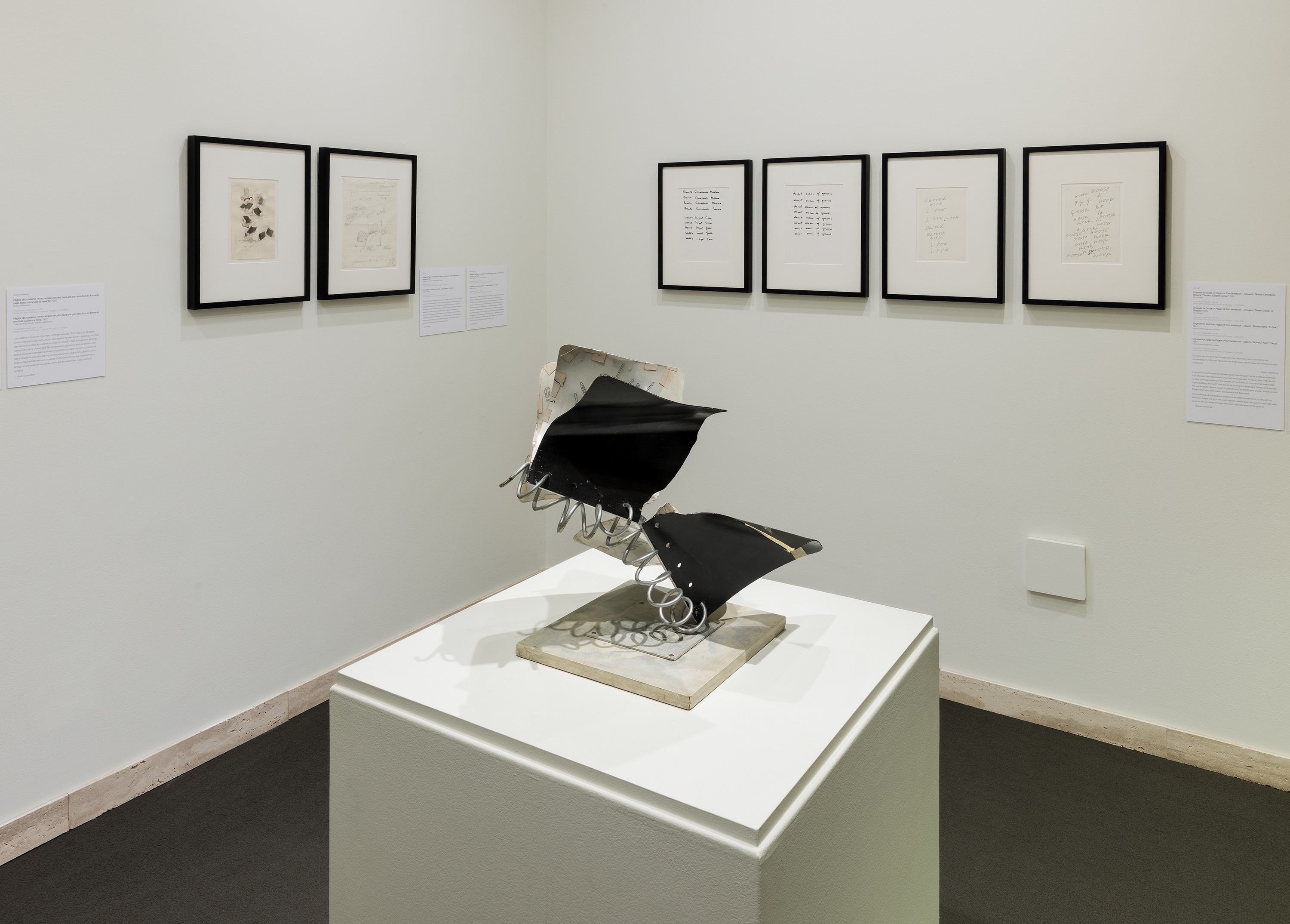 Modern sculpture displayed on a pedestal in an art gallery, surrounded by framed documents on walls.