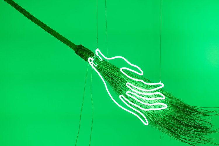 Glowing white neon hand on a broomstick against vibrant green background.
