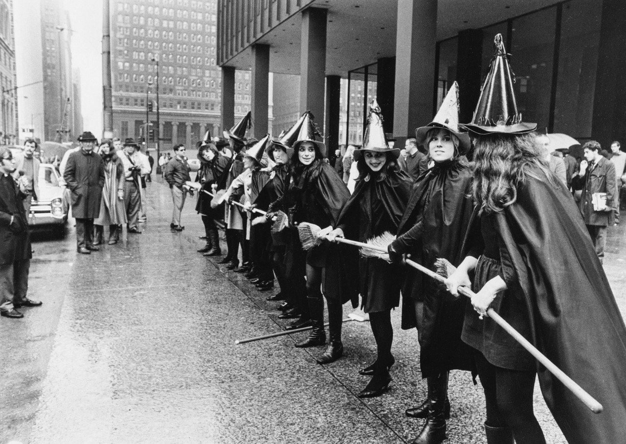 Women in witch costumes, lined up on a city street, holding broomsticks, with a crowd nearby.