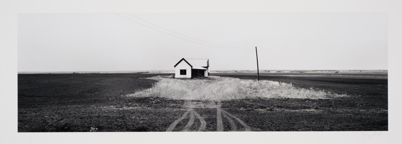 A small white house stands isolated in a flat, barren field with a dirt path leading to it.