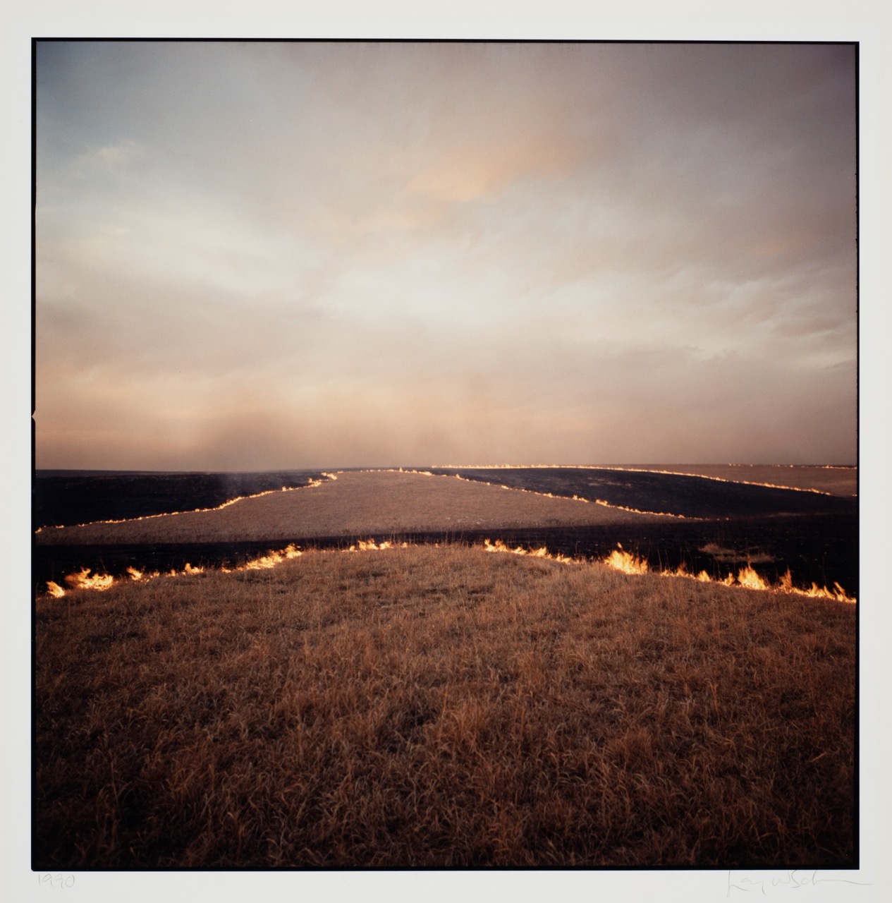 Burning grass fields under a cloudy sky at dusk.