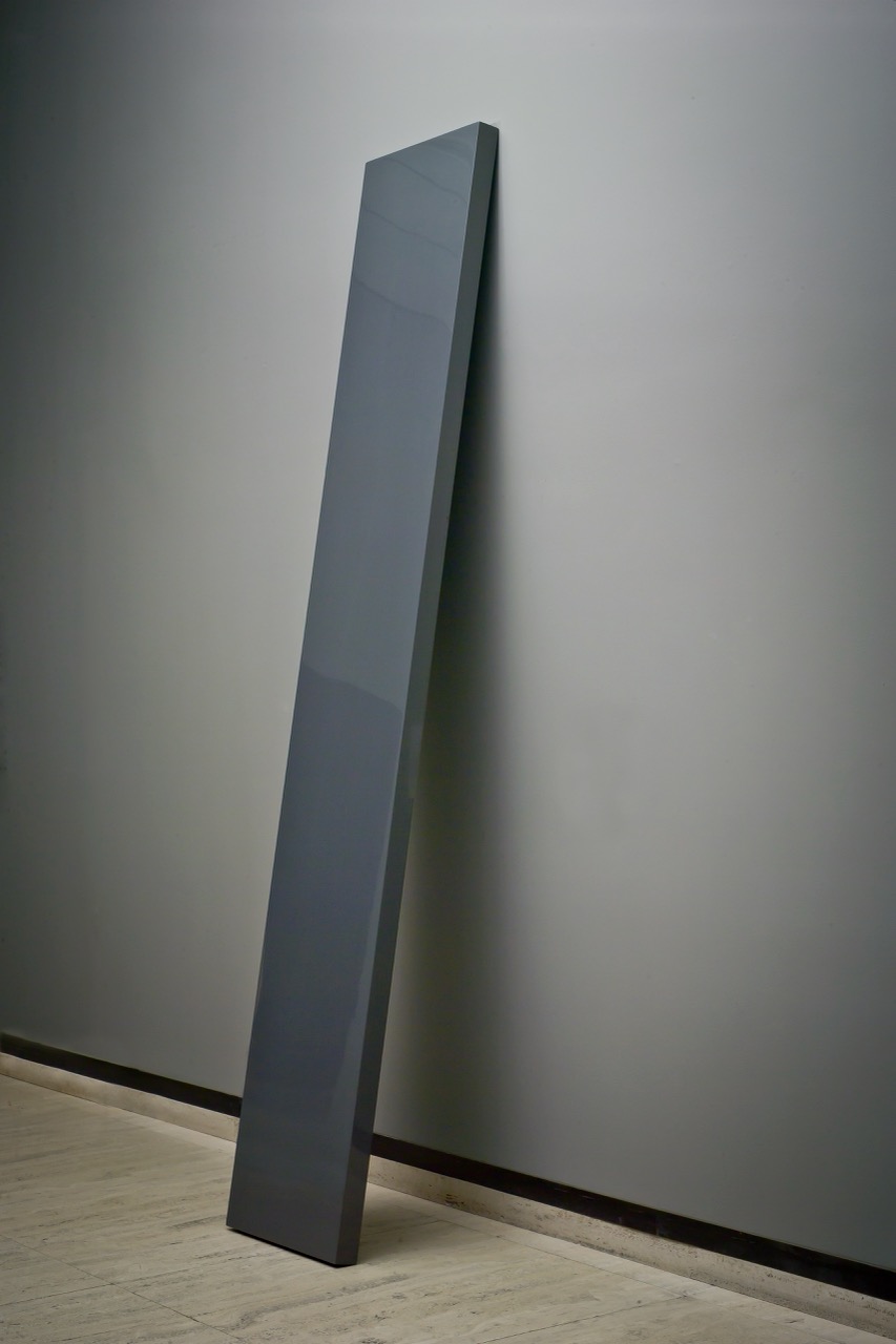Tall, dark gray rectangular sculpture leaning against a light gray wall on a travertine floor.