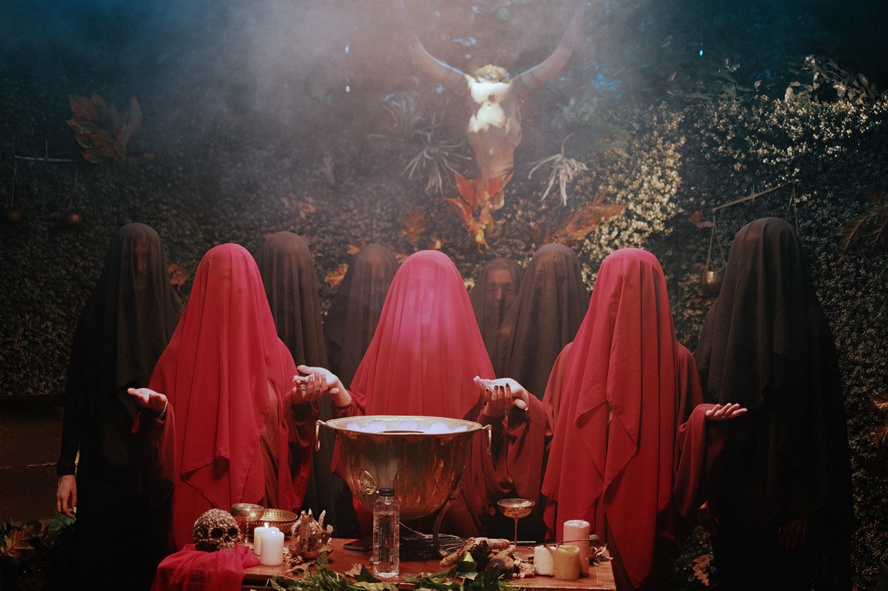Figures in red and black veils surround a silver bowl in a mystical, forested ritual setting.