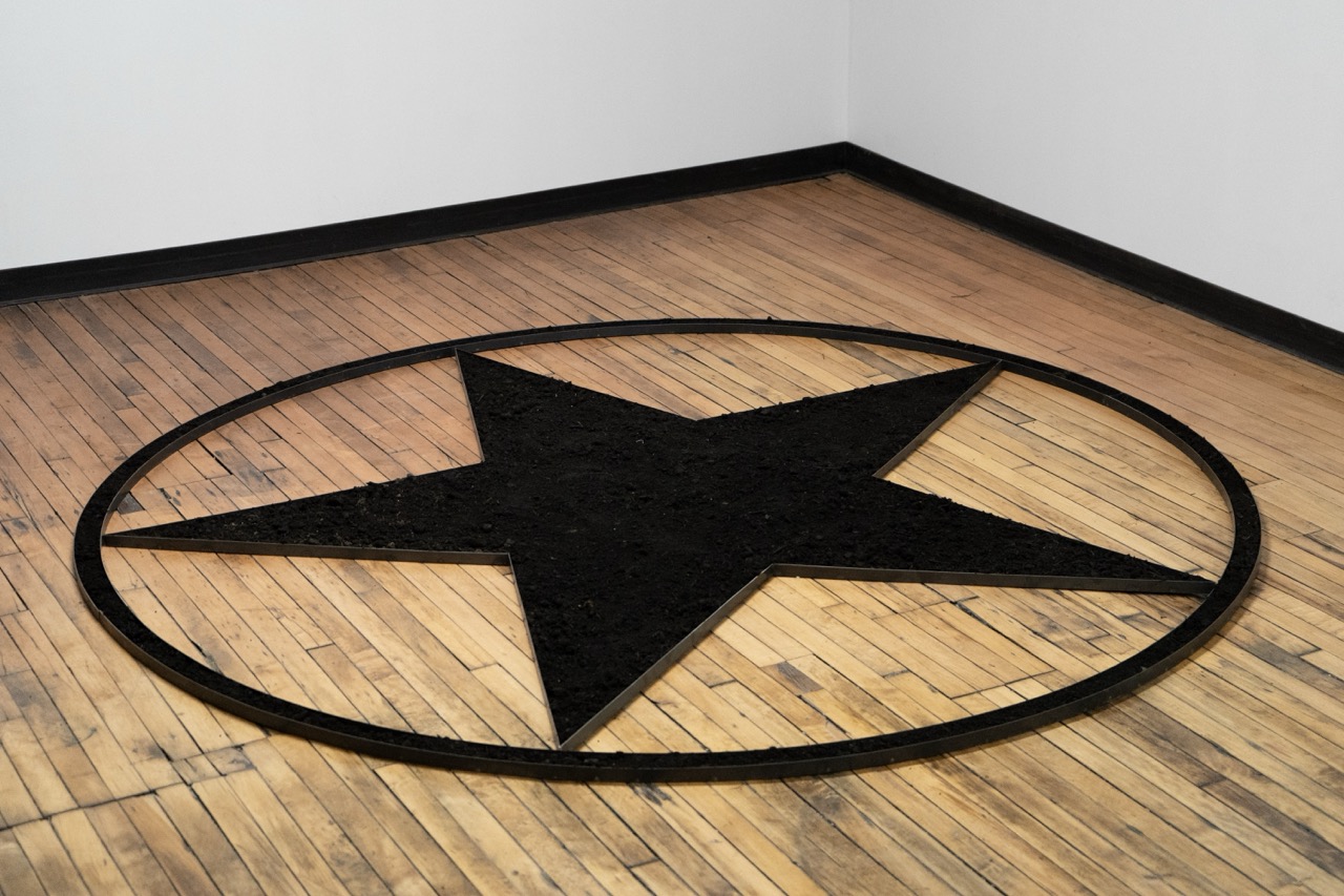 Black star artwork on a wooden floor, enclosed in a circular frame, situated in a minimalist room.