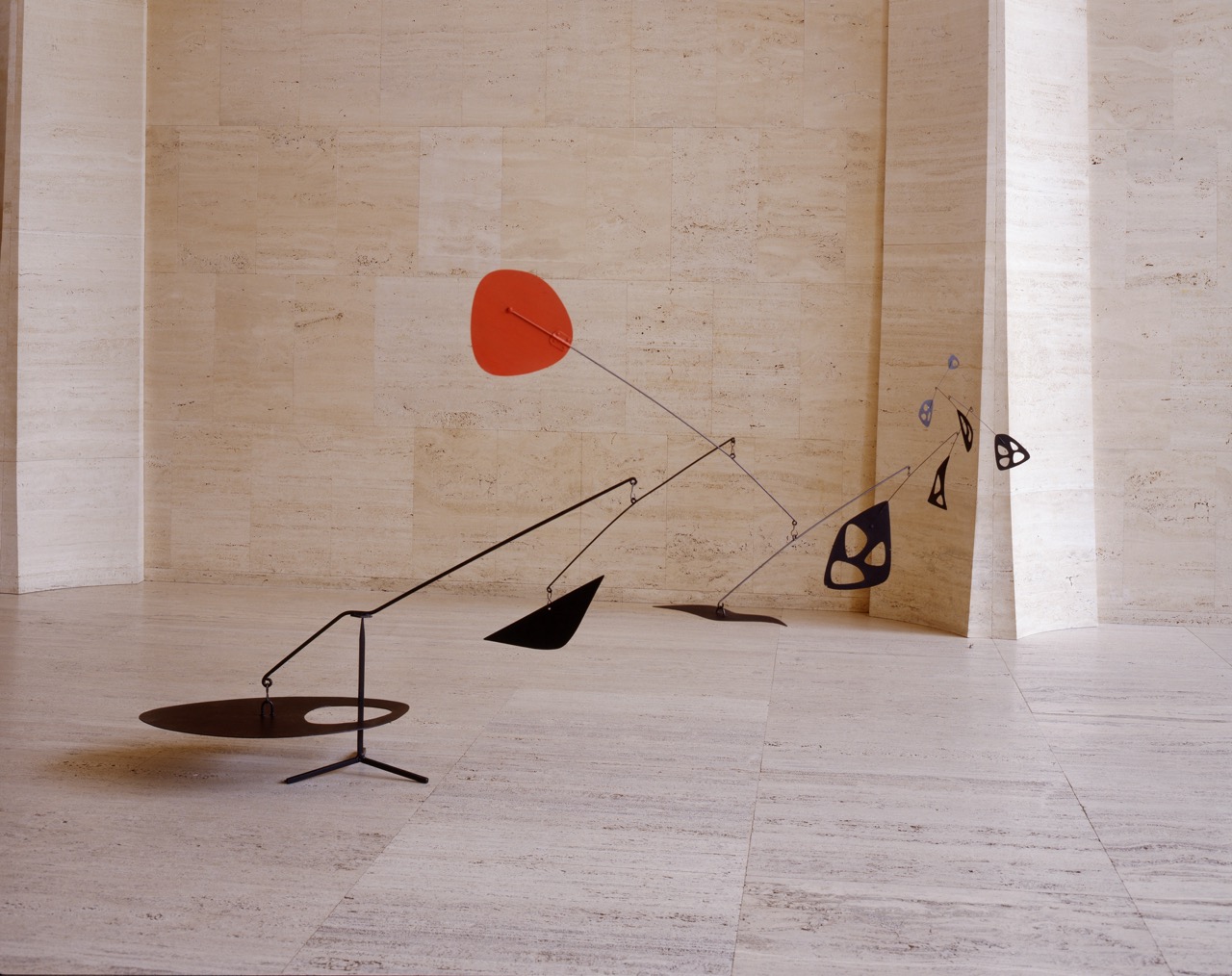 Mobile sculpture with red, black, and blue abstract shapes in a beige stone room.