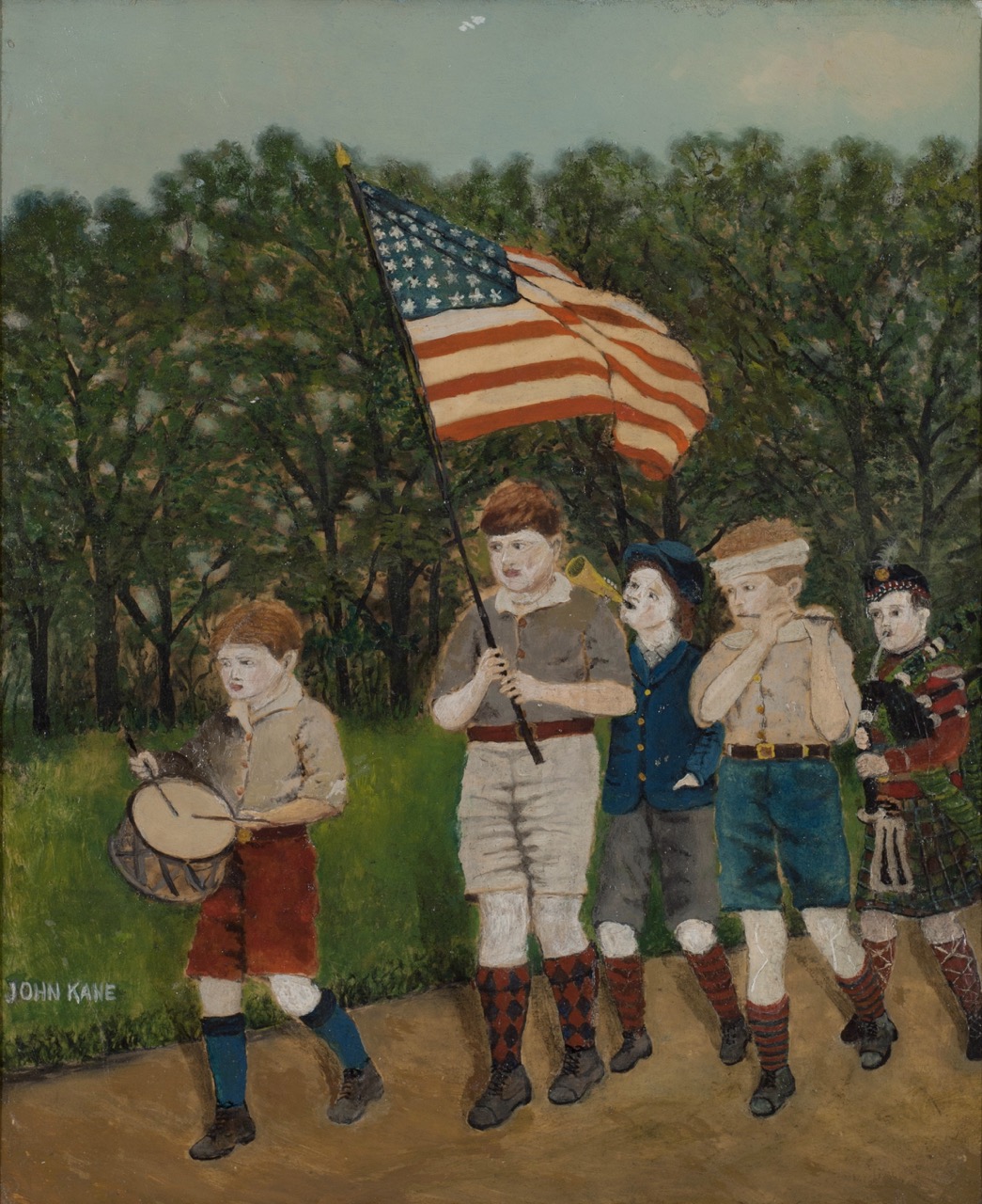 Children march on a dirt path holding a U.S. flag, drum, and dressed in festival attire.