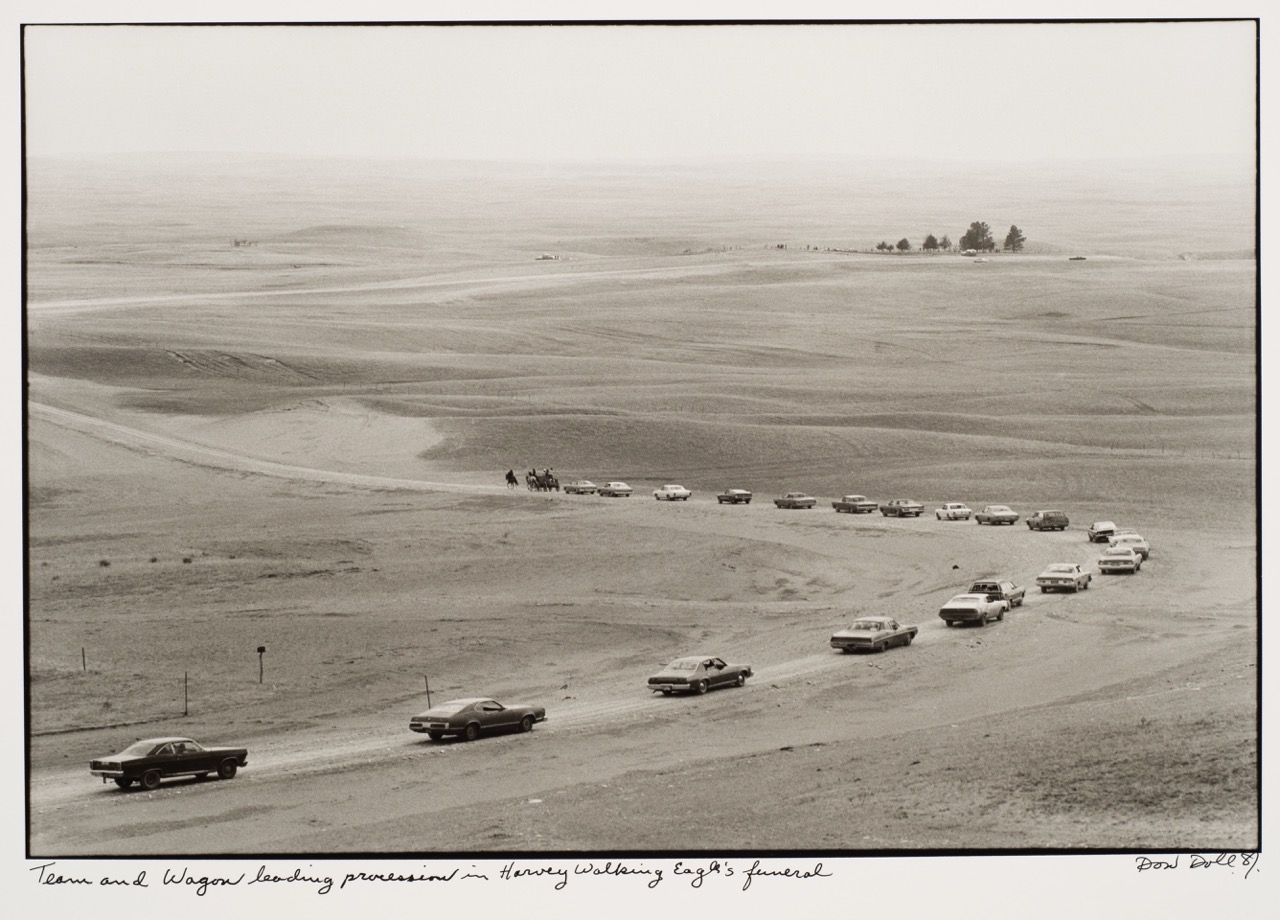 A line of cars drives through a vast, open landscape, followed by horseback riders in the distance.