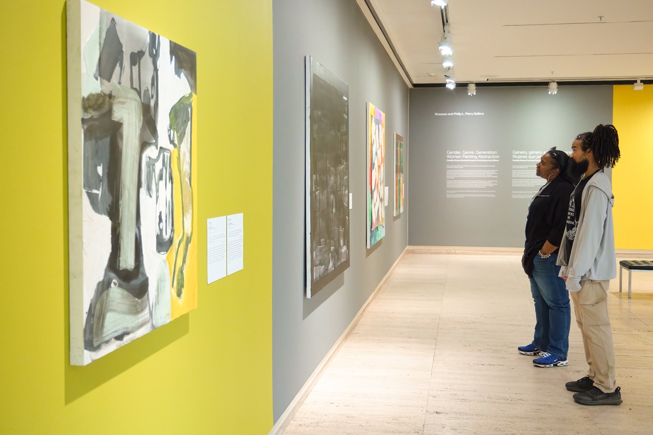 People admiring colorful paintings in a modern art gallery with yellow and gray walls.
