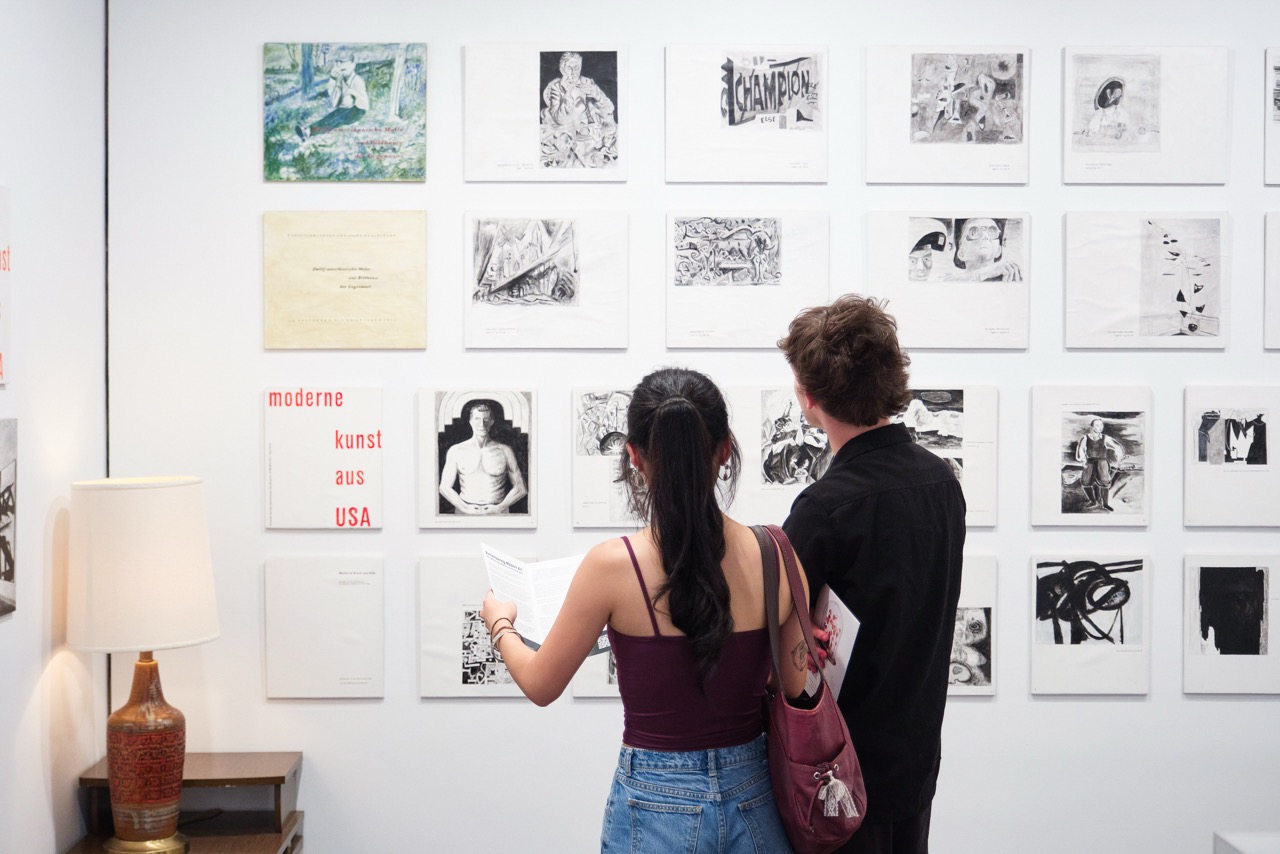 Two people view a wall displaying monochrome art and posters in a gallery setting.