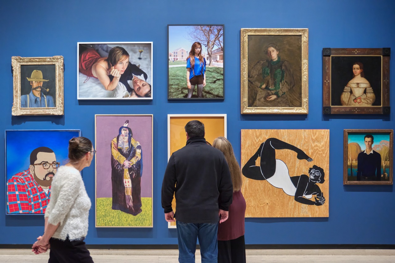 People viewing a gallery wall featuring various portraits and artwork on a blue background.