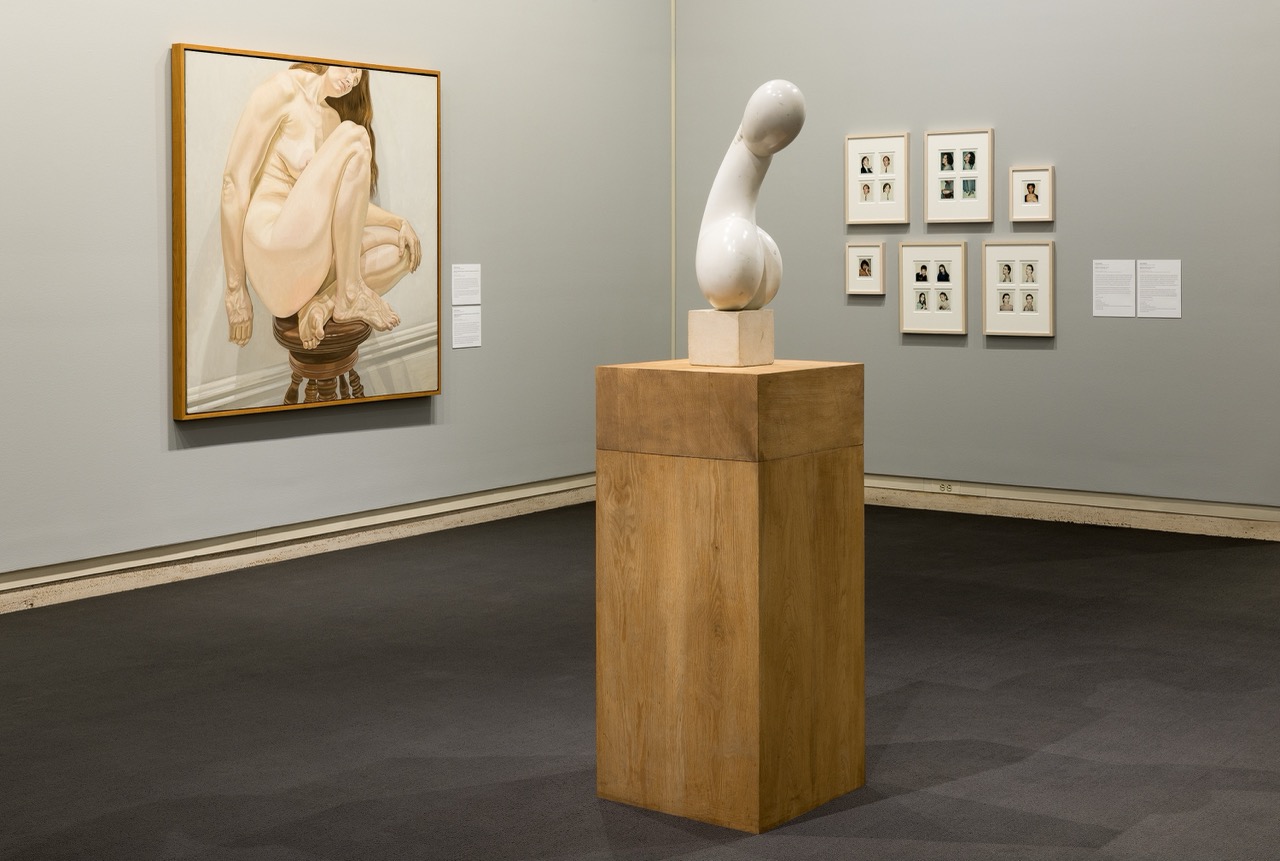 Gallery interior with abstract sculpture on a wooden pedestal and framed art pieces on the walls.